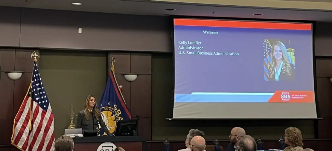 Kelly Loeffler, Administrator of U.S. Small Business Administration, speaking at a podium during a presentation, with U.S. and SBA flags displayed beside her. A large screen shows a welcome slide.