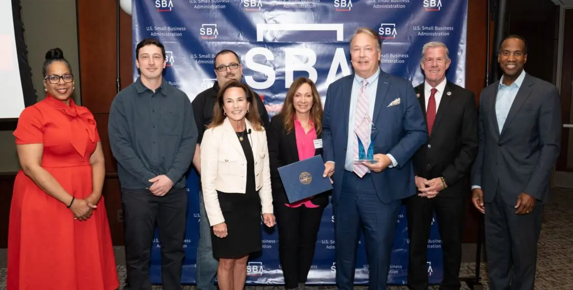 Bruce Barron in a blue suit holds a trophy and a certificate, surrounded by 7 other formally dressed Barron team members standing in front of a blue backdrop with U.S. Small Business Administration logos at the SBA event