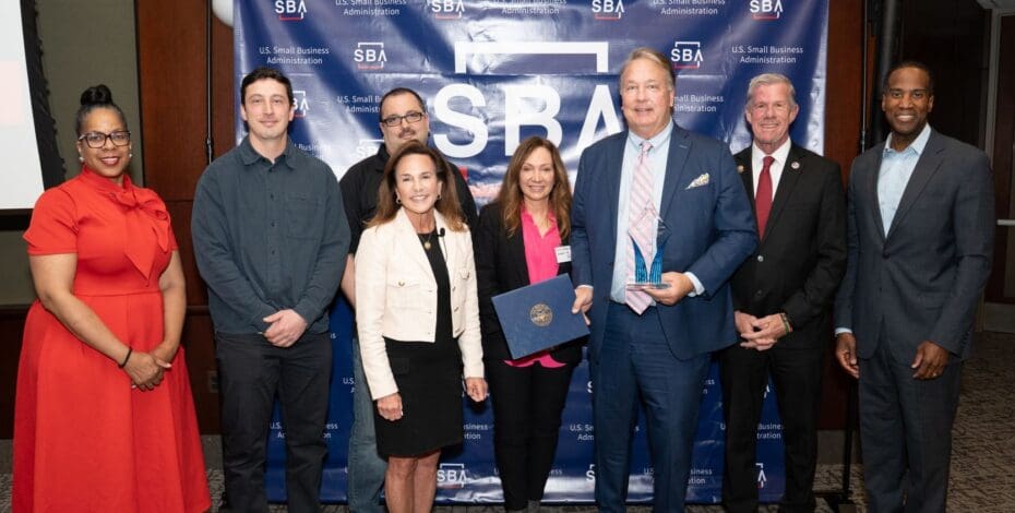 Bruce Barron in a blue suit holds a trophy and a certificate, surrounded by 7 other formally dressed Barron team members standing in front of a blue backdrop with U.S. Small Business Administration logos at the SBA event